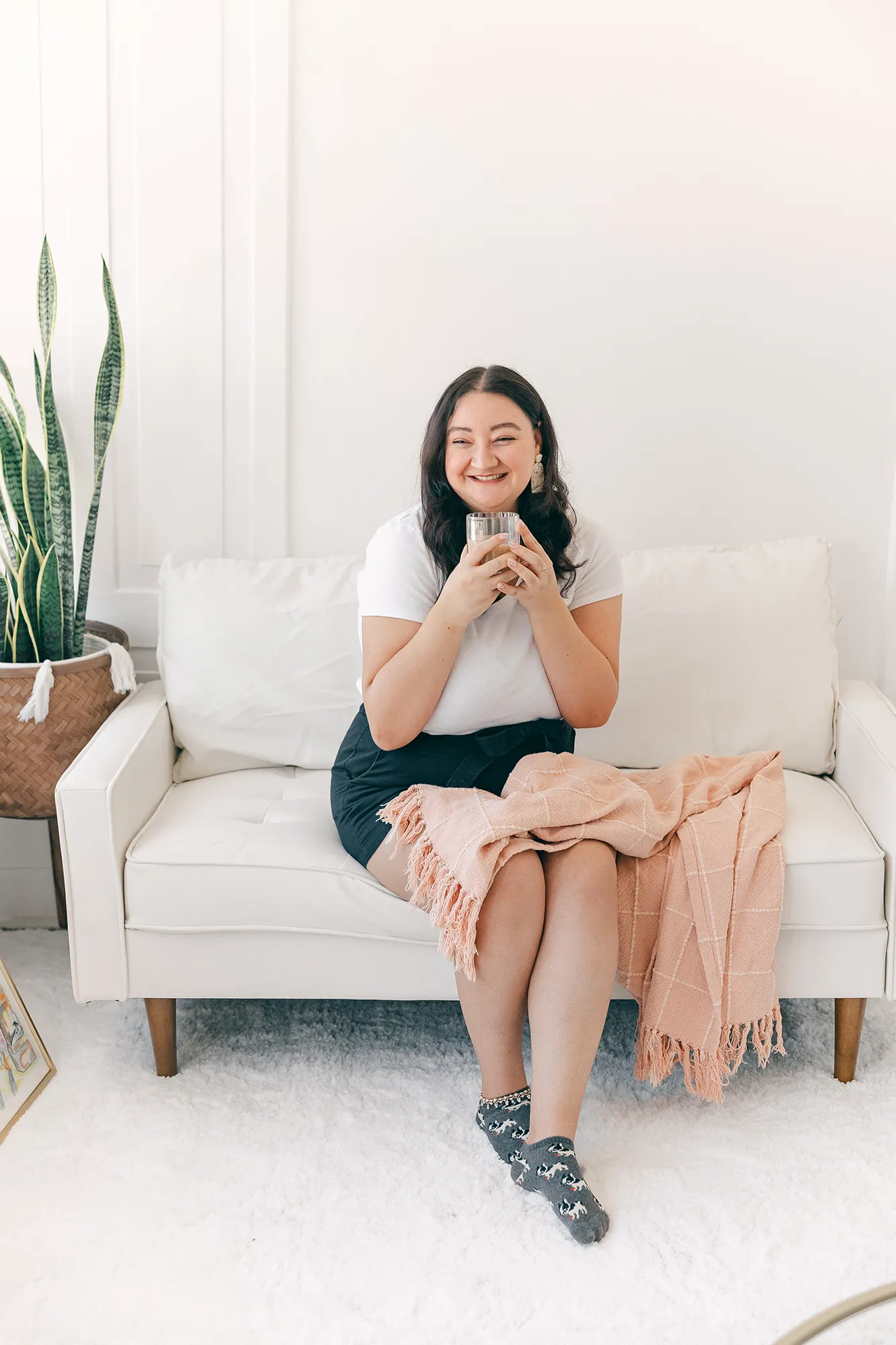 Kimberly sitting on a white couch with a pink throw blanket, smiling and drinking tea